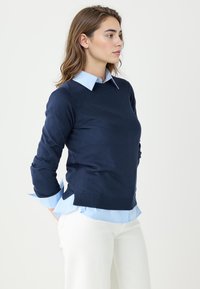 Granatowy sweter z długimi rękawami, noszony na jasnoniebieskiej koszuli w paski. Sweter ma klasyczny okrągły dekolt i ściągacz na dole.