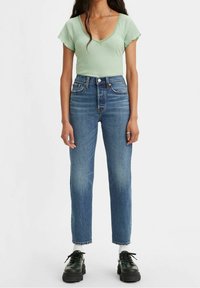 Mujer viste una blusa de manga corta verde claro, jeans de tiro alto azul, calcetines blancos y zapatos negros gruesos, de pie contra un fondo blanco.