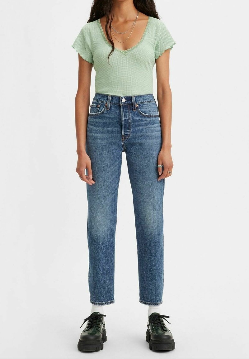 Mujer viste una blusa de manga corta verde claro, jeans de tiro alto azul, calcetines blancos y zapatos negros gruesos, de pie contra un fondo blanco.