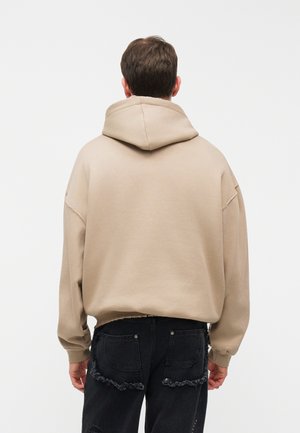 Homme portant un sweat à capuche oversize beige et un jean noir déchiré, debout de dos sur un fond blanc uni.