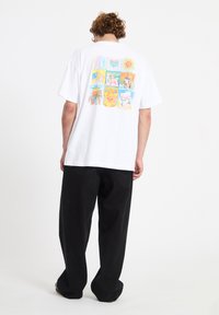 Άτομο με σγουρά μαλλιά που φοράει λευκό φαρδύ T-shirt με πολύχρωμα σχέδια εννέα πάνελ και χαλαρό μαύρο παντελόνι, στέκεται με την πλάτη προς το μέρος μας.