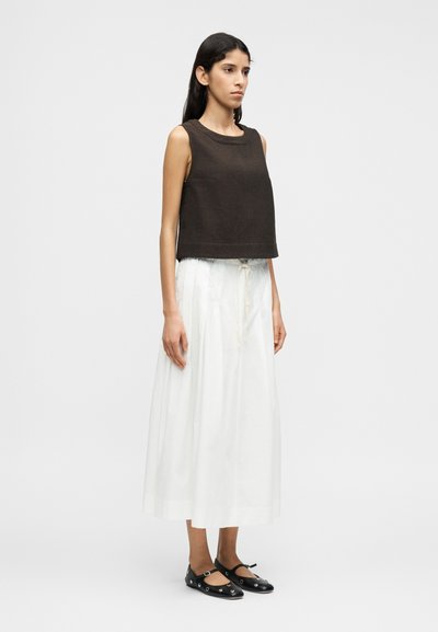 Καφέ κοντό τοπ χωρίς μανίκια, σε συνδυασμό με μακριά λευκή φούστα με κορδόνι στη μέση. Μαύρα σανδάλια με επίπεδο πάτο και κυκλικές λεπτομέρειες.