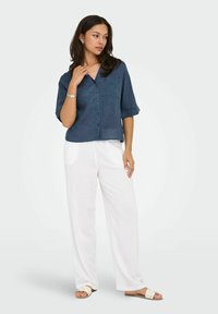 Camicia button-up in lino blu navy a maniche corte con scollatura a V e taschino sul petto, abbinata a pantaloni larghi bianchi e sandali chiari.