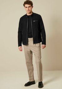 Chaqueta negra con cremallera y cuello alzado, acompañada de pantalones beige claro y zapatos de vestir negros. Textura de tela suave.