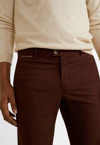 Brązowe spodnie wykonane z bawełny o gładkiej fakturze, z zamkiem błyskawicznym z przodu i zapięciem na guzik, z dyskretnymi detalami bocznych kieszeni.
