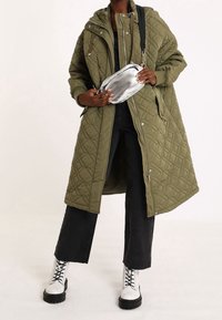 Olijfgroene gewatteerde lange jas met capuchon, gecombineerd met zwarte flared jeans en witte veterschoenen. Een zilveren crossbody tas voegt contrast toe.