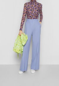 Długie rękawy, bluzka w kwiatowy wzór w odcieniu fioletowym, spód spodni w jasnoniebieskim kolorze z wysokim stanem, trzymająca zieloną kurtkę z efektem tie-dye, dopełniona białymi sneakersami.