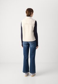 Fleece vest in cream, collared, sleeveless, worn over a navy long-sleeve top, paired with flared blue jeans and light-colored shoes.