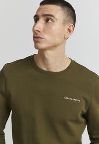 Olivengrønn sweatshirt med rund hals og ribbede mansjetter, med hvit tekst "CASUAL FRIDAY" på venstre bryst. Glatt stoffstruktur.