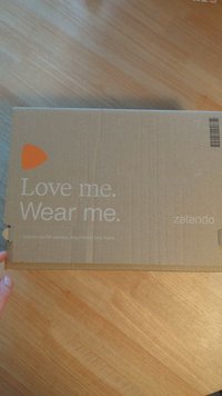 Hnědá kartonová krabice s horním panelem, na kterém je velké oranžové logo a bíly text: "Miluj mě. Nosi mě. Rozbal mě pro příběhy, inspiraci a další."