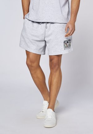Mann trägt hellgraue Sportshorts mit buntem "Since 1982 Active Wear"-Logo am linken Oberschenkel und weißen Turnschuhen.