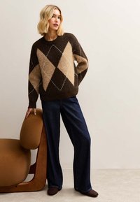 Pull à losanges marron et beige avec une coupe décontractée, associé à un jean bleu foncé à jambes larges. Une chaise ronde marron se trouve en arrière-plan.