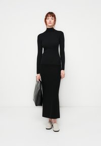 Svart, rillet turtleneck genser kombinert med et svart langt skjørt. Modellen bærer en svart håndveske. Hun har på seg lysfargede slip-on sko.