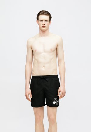 Νεαρός άνδρας στέκεται μπροστά σε απλό φόντο φορώντας μαύρο μαγιό κολύμβησης Nike με λευκό λογότυπο, χωρίς μπλουζάκι και κοιτάζοντας μπροστά.