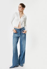 Weißes, strukturiertes, langärmeliges cropped Shirt kombiniert mit hochtaillierten, weit geschnittenen blauen Jeans. Das Model trägt spitz zulaufende graue Schuhe.