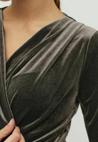 Robe en velours gris foncé, présentant un design portefeuille avec un drapé doux et une texture lisse. Manches longues et léger éclat qui rehaussent son apparence.