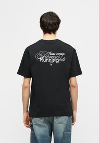 Άτομο που φαίνεται από πίσω, φοράει μαύρο μπλουζάκι με λευκό κείμενο που λέει "ημερήσια άσκηση: Πάντα αργοπορημένος" και μπλε τζιν.