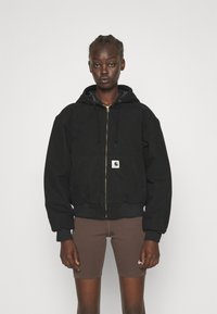 Black zip-up hoodie with side pockets, ribbed cuffs and hem, gold zipper, and logo patch. Worn over brown bike shorts.
