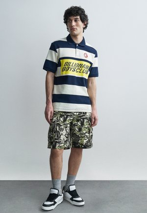 Jaunas vyras dėvi tamsiai mėlyną ir baltą dryžuotą polo marškinėlį su užrašu „Billionaire Boys Club“, kamufliažo krovinių šortus, pilkas kojines ir juodai baltus sportbačius.