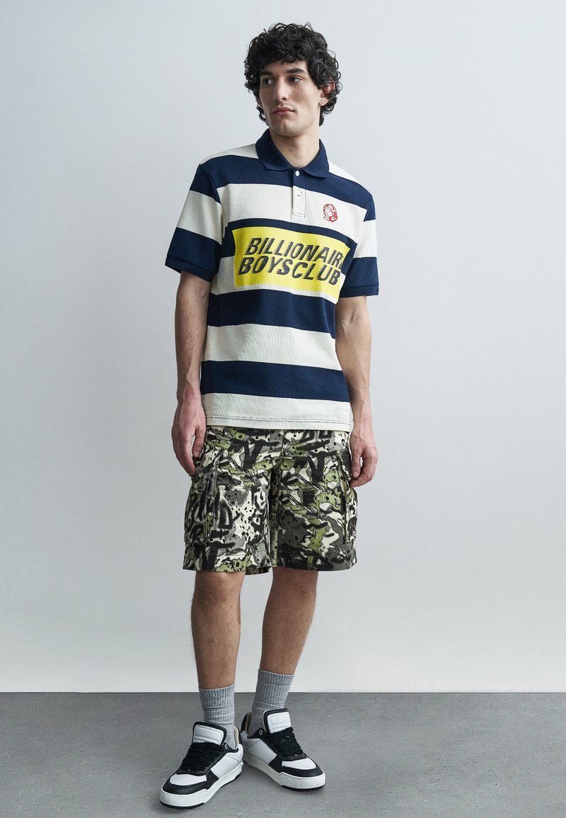 Νεαρός άνδρας που φοράει μπλε σκούρο και λευκό ριγέ πόλο με το κείμενο "Billionaire Boys Club", καμουφλαρισμένη κοντή βερμούδα cargo, γκρι κάλτσες και ασπρόμαυρα αθλητικά παπούτσια.