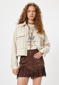 Chaqueta recortada de color beige claro con grandes bolsillos, superpuesta a una camiseta gráfica de color crema. Minifalda de ante marrón con detalles de flecos.