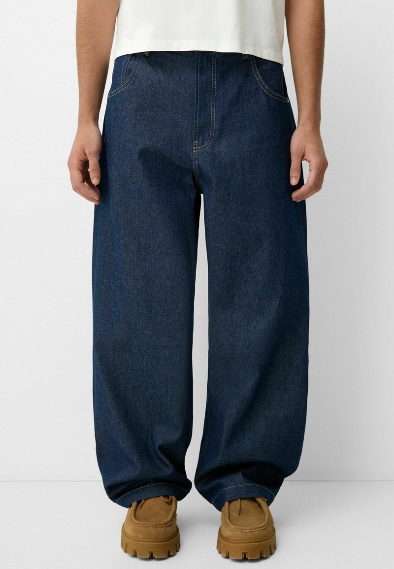 Løse mørke denimjeans med synlige syninger, store forlommer og lige ben. Kombineret med brune støvler mod en neutral baggrund.