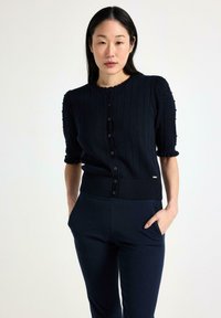 Cardigan navy a costine con maniche corte a sbuffo, chiusura con bottoni e dettagli testurizzati sulle maniche. Indossato con pantaloni scuri abbinati.
