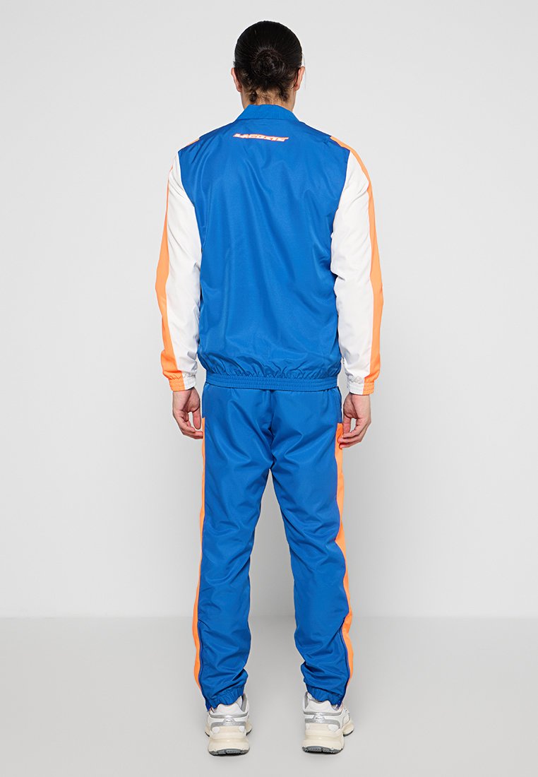 Chándal azul con rayas naranjas en las mangas y los pantalones, hecho de una tela suave. Puños y cintura elásticos, con un detalle de logo en la parte posterior.