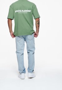 Osoba viděná zezadu, oblečená v zeleném tričku Pegador, světlých modrých džínách, bílých teniskách a s tetováním na pažích, na bílém pozadí.