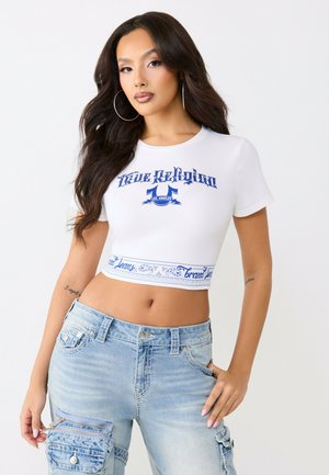Jeune femme aux longs cheveux foncés portant des boucles d'oreilles créoles, un crop top blanc avec un texte bleu, et un pantalon cargo en denim bleu clair avec une petite pochette.