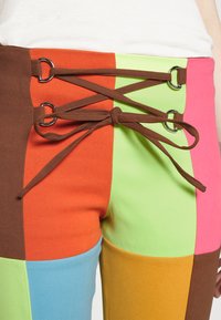 Calças com design em patchwork colorido, com secções em verde, laranja, rosa, azul e castanho. Apresentam um cordão castanho passado por ilhós de metal prateado.