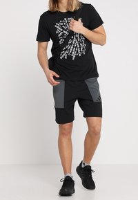 Mustapaita, jossa on valkoista tekstiä, ja musta-harmaat urheilushortsit, joissa on kulmikas värierottelu. Mustat urheilujalkineet viimeistelevät lookin.