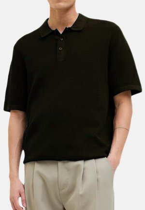 Schwarzes Poloshirt aus strukturiertem Stoff, mit kurzen Ärmeln und einer Knopfleiste mit drei Knöpfen. Kombiniert mit hellen Hosen.