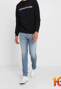 Felpa nera con la scritta "CALVIN KLEIN JEANS" in bianco, jeans azzurri e sneakers bianche con dettagli rossi. Look casual.