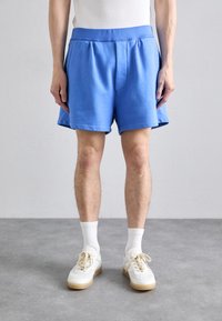 Pantalones cortos de algodón azul claro con cintura elástica, pliegue frontal y bolsillos laterales, combinados con zapatillas blancas y calcetines blancos de longitud media.