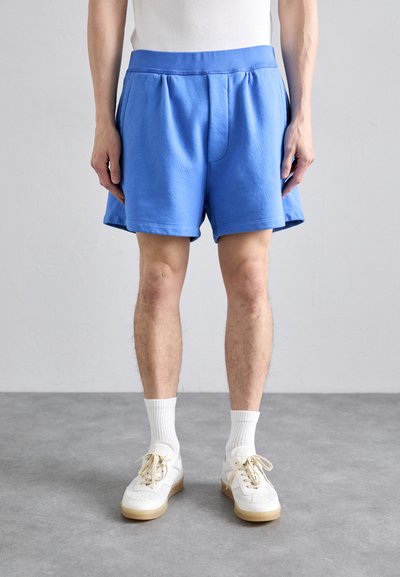 Pantaloni corti in cotone azzurro chiaro con cintura elastica, piega frontale e tasche laterali, abbinati a sneakers bianche e calzettoni bianchi di lunghezza media.