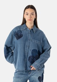 Blå denimskjorte med store mørkeblå paljett-hjertepatcher. Har knappelukking foran, krage og brystlomme; uferdig kant nederst.