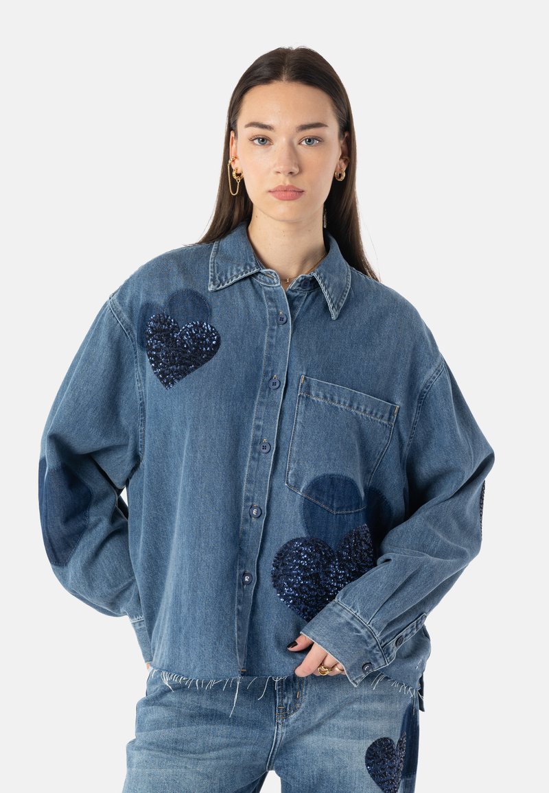 Blå denimskjorte med store mørkeblå paljett-hjertepatcher. Har knappelukking foran, krage og brystlomme; uferdig kant nederst.