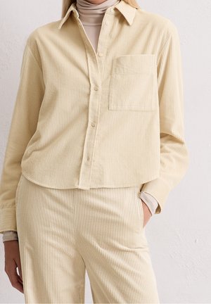 Beige corduroy button-up shirt with chest pocket, paired with matching corduroy pants and a beige turtleneck underneath.