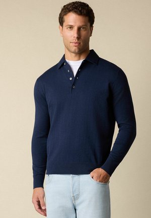 Námořnická polo košile s dlouhým rukávem, límečkem, třemi knoflíky a žebrovanými manžetami, kombinovaná se světle modrými džíny. Hladká textura.
