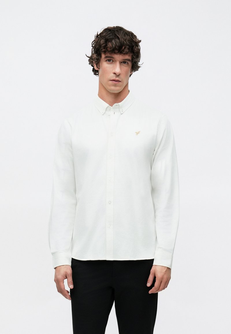 Biela košeľa s dlhým rukávom a gombíkmi, vyrobená z hladkej látky, so malým zlatým logom na ľavej strane na hrudi a tradičným golierom.