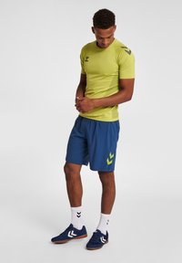 Lichtgroen sportshirt met korte mouwen en mesh zijpanelen, gecombineerd met donkerblauwe shorts met een lichtgroen chevronlogo.