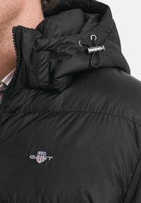 Zwarte pufferjack met een ritskap met trekkoord, een gladde textuur en een geborduurd logo op de borst. Compact ontwerp.