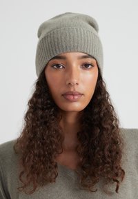 Joven mujer con cabello rizado largo, usando un gorro de punto gris y un suéter a juego, mirando directamente a la cámara con una expresión neutral.