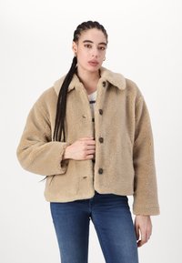Beige sünteetilisest karusnahast jakk, millel on kaelus, nupud ja pikad varrukad. Tekstuur näib olevat pehme ja luksuslik.