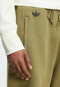 Olivově zelené tepláky se bočními kapsami, stahovací pas a vyšívaným námořnickým modrým logem na stehně. Textura z bavlněné směsi.