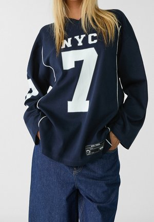Personne portant une chemise bleu marine à manches longues avec "NYC 7" et un jean bleu décontracté, les mains dans les poches, sur un fond uni clair.