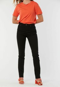 T-shirt en coton orange assortie à un jean skinny noir. Le modèle porte des sandales à talons carrés rouges. Design simple, sans motifs, textures lisses.