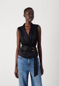 Black satin sleeveless top with a wrap design, featuring a deep V-neck and a waist tie with a gold buckle. Paired with light blue jeans.