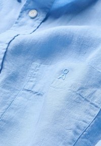 Gros plan sur une chemise en lin bleu clair avec coutures visibles, bouton et logo brodé au-dessus de la poche poitrine.